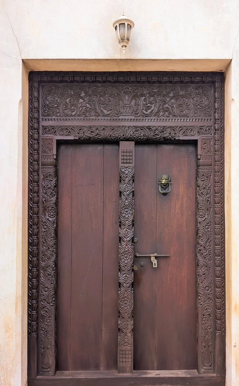 A dark hand-carved Lamu double door with a floral frame, a carved centre post and a brass lion-head knocker