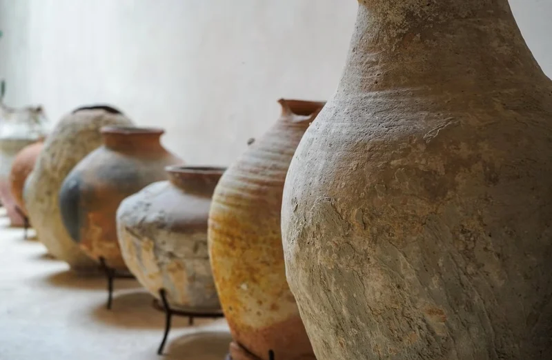 A row of weathered clay jars on iron stands against a lime-washed wall, the nearest one tall and narrow-necked