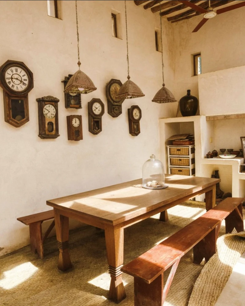 A limewashed coastal dining room: a wooden table and benches, a wall of antique clocks and woven pendant lamps