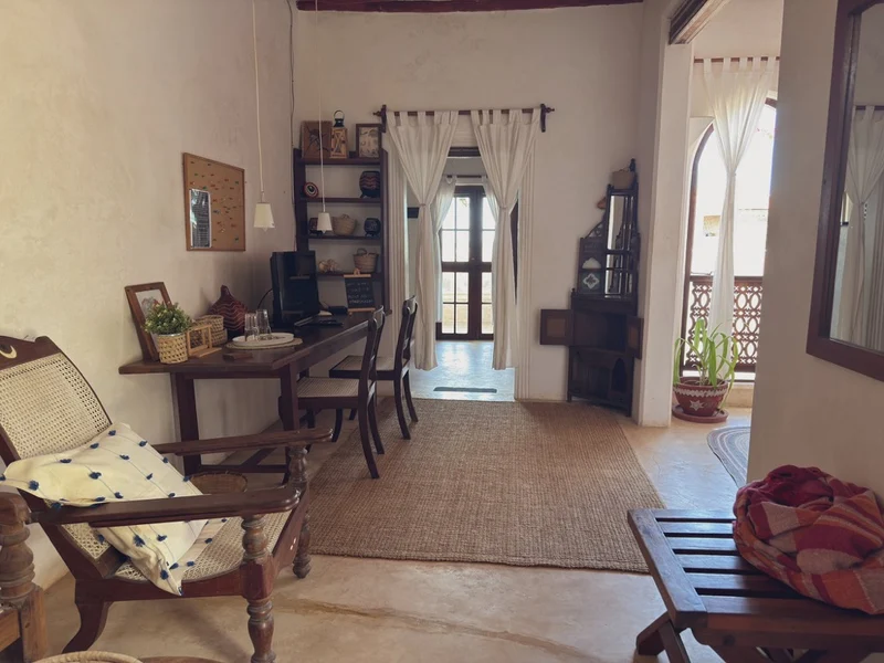 A coastal room with a planter's chair, a writing desk, carved shelves and an arched door to the balcony