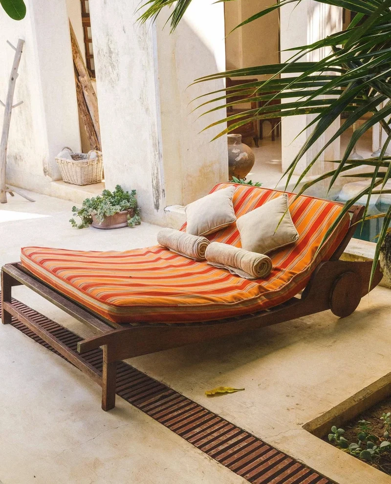 A wooden day bed with striped cushions and bolsters in a sunlit courtyard under palm fronds