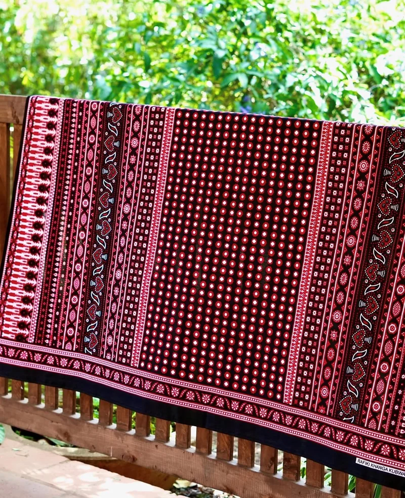 A red and black kanga with patterned borders, draped over a wooden balcony rail