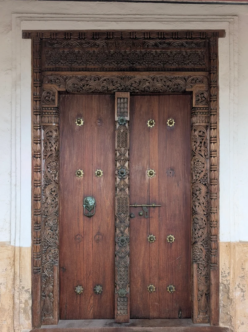 A hand-carved Lamu double door: floral and scroll carving on the frame, a carved centre post, brass studs and a lion-head knocker