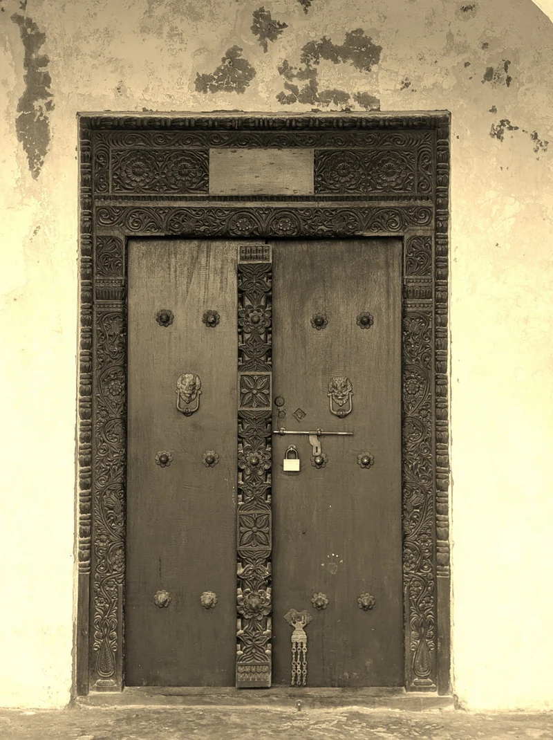 An old carved Swahili door with brass studs, two lion-head knockers and a chained hasp, in sepia