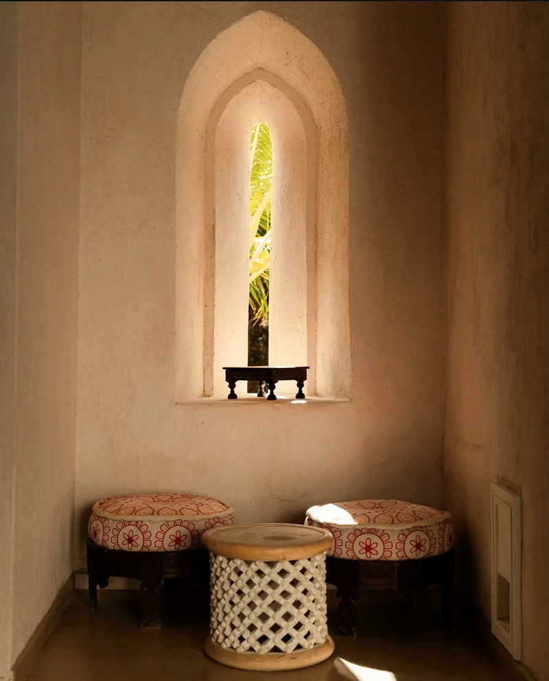 A limewashed alcove with an arched window niche, two cushioned stools and a carved side table