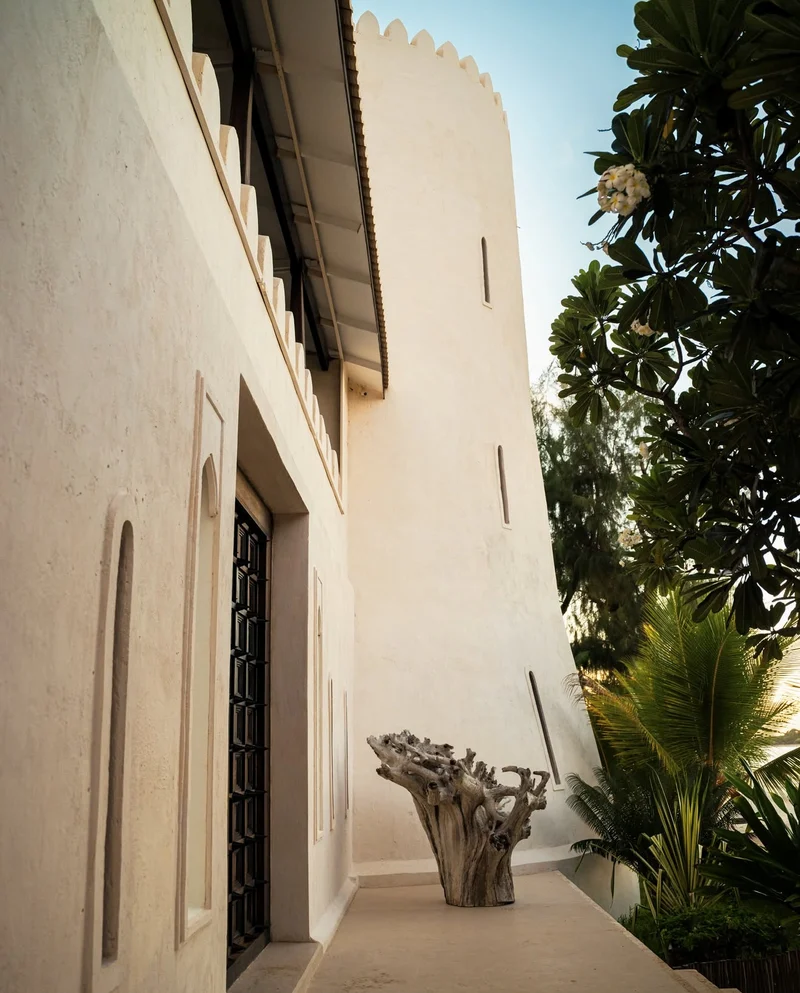 A narrow whitewashed passage under crenellated walls, with a driftwood sculpture and palms