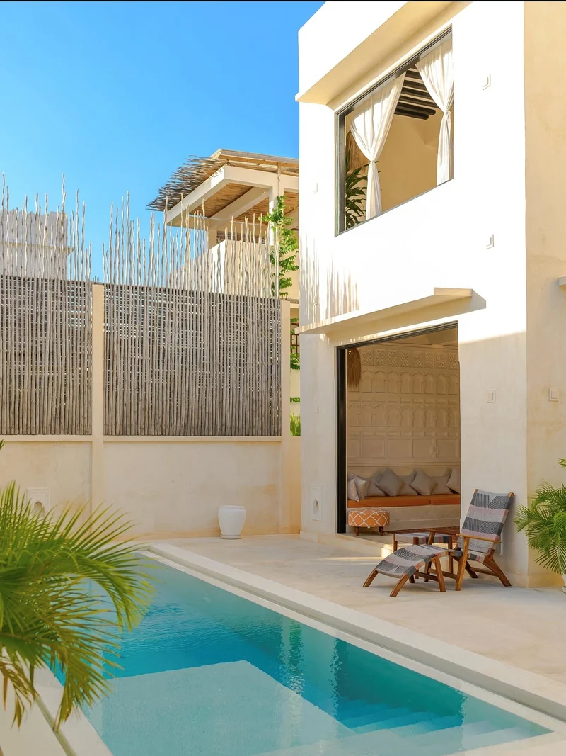 A pool terrace with a timber deck chair before open doors to a cushioned sitting room
